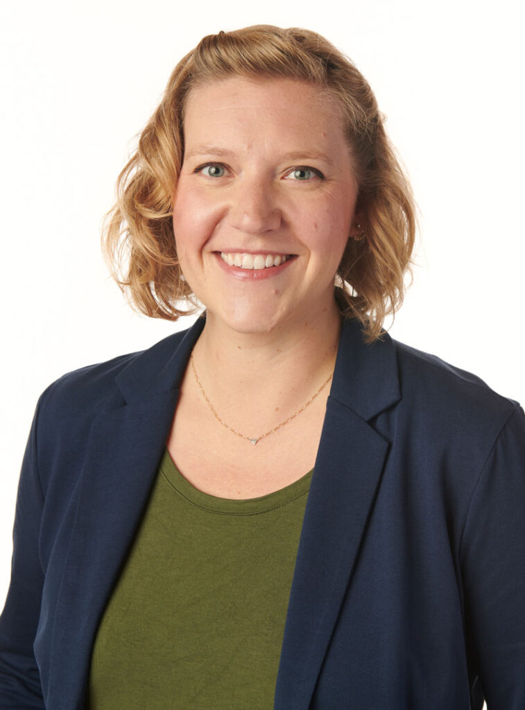 headshot of sarah jerger smiling wearing a navy blue blazer over a hunter green shirt